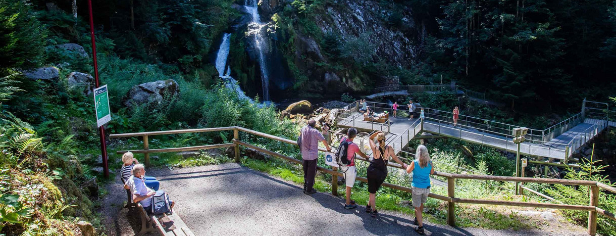Touristen stehen an einem Geländer und auf einer Aussichtsplattform und blicken auf eine Stufe der Triberger Wasserfälle, über die Wasser fließt.